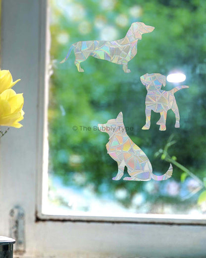 The three dog-shaped sun catchers arranged vertically on a white-framed window. A soft blur of greenery is visible through the glass in the background.