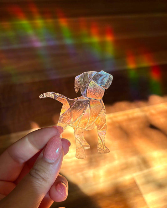 Hand holding a transparent dog-shaped sun catcher sticker with a background of rainbow light rays.