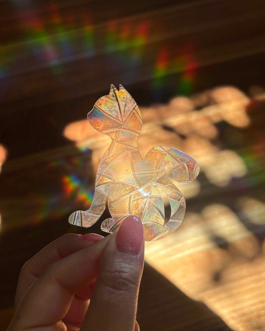 Iridescent cat-shaped sun catcher sticker held by a hand with a blurred rainbow lights background