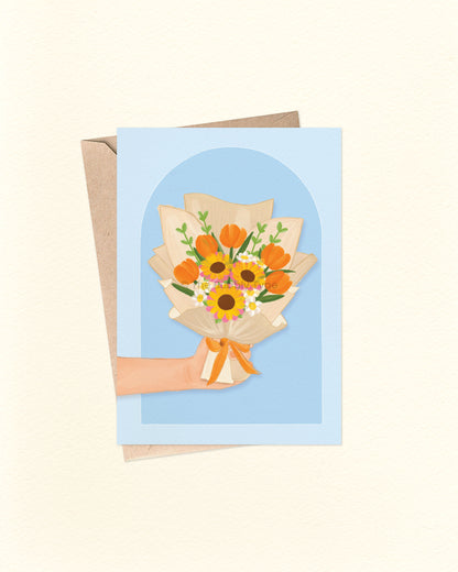 Front of A6 greeting card featuring an illustration of a hand holding a bouquet of yellow and orange flowers with green foliage, wrapped in creamy white paper. Set against a blue arch-shaped background. A versatile Australian made card by The Bubbly Type – perfect for everyday messages like thank you, thinking of you, or just because. Shown with a brown kraft envelope.