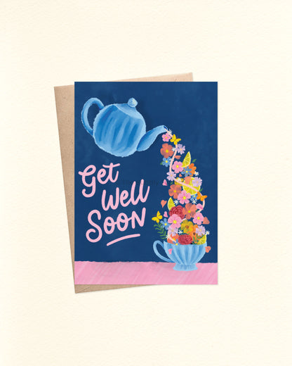 A6 greeting card with a rich deep blue background. The illustration shows a watercolour-style blue teapot tilted and pouring out a flow of yellow lemons, pink and orange flowers, green foliage, and butterflies into a matching blue teacup. To the right, in pink hand-lettered script, it reads “get well soon.” Shown with a brown kraft envelope behind the card. Used to give well wishes to people with sickness who need rest and rejuvenation. 