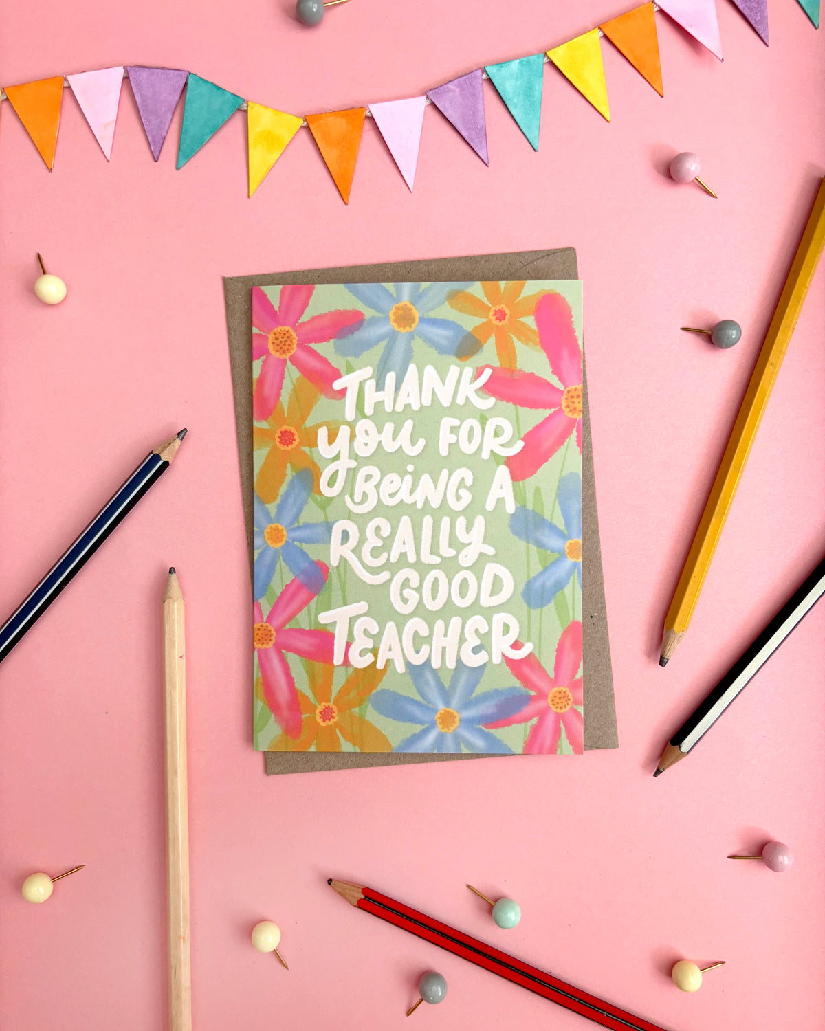 A square thank-you card with a floral design and the text 'THANK YOU FOR BEING A REALLY GOOD TEACHER' in white lettering, placed on a pink background with pencils, beads, and bunting.