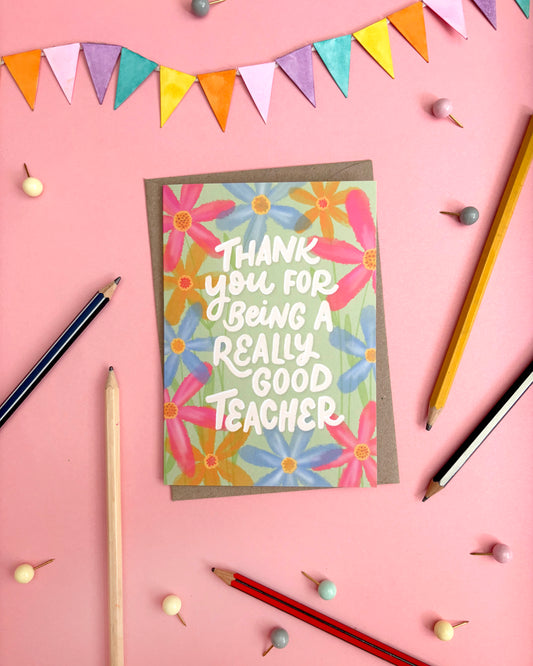 A square thank-you card with a floral design and the text 'THANK YOU FOR BEING A REALLY GOOD TEACHER' in white lettering, placed on a pink background with pencils, beads, and bunting.