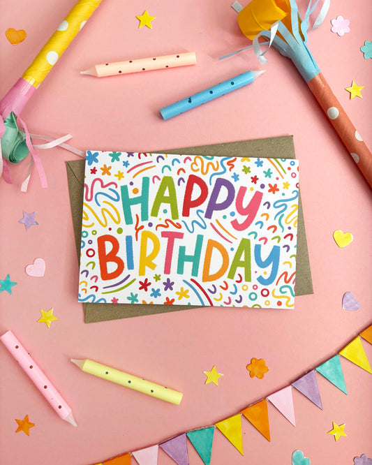 A colourful 'Happy Birthday' greeting card with playful multicoloured lettering and doodles, placed on a brown envelope on a pink background. The scene is styled with party props including candles, party hats, star and heart confetti, and bunting.