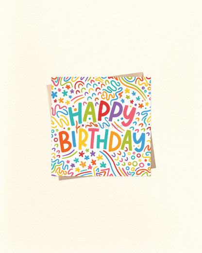 A square birthday card with a white background covered in colourful doodle patterns and the words 'HAPPY BIRTHDAY' in playful multicoloured lettering, shown on a warm off-white background.