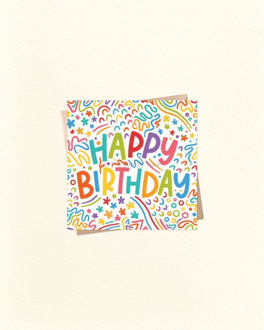 A square birthday card with a white background covered in colourful doodle patterns and the words 'HAPPY BIRTHDAY' in playful multicoloured lettering, shown on a warm off-white background.