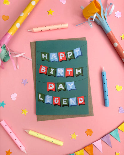 A handmade birthday card reading 'HAPPY BIRTH DAY LEGEND' in colourful letter flags, displayed on a pink background with party props like candles, streamers, and star decorations.