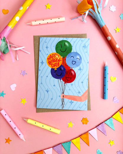 A birthday card featuring a hand holding a cluster of colourful smiley-face balloons, placed on a pink surface with party props like candles, confetti, and bunting.
