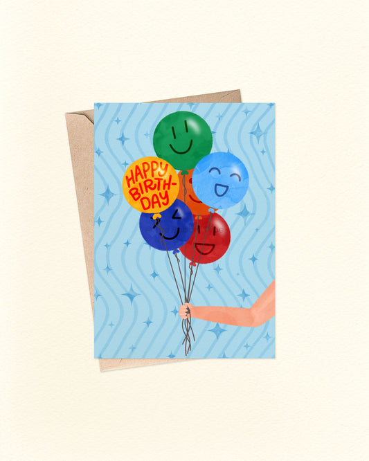 Front of A6 birthday card with a cheerful illustration of a hand holding a bunch of colourful balloons—green, blue, dark blue, red, and yellow. Each balloon has a smiley face, except the yellow one which reads “Happy Birthday.” The blue background features playful wavy lines and star accents. Displayed with a brown kraft envelope.