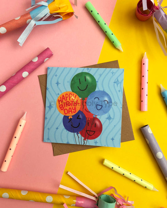 A square birthday card with colourful smiley-face balloons illustrated on a groovy blue background, placed on a brown envelope over a pink and yellow background. Craft paper party horns and candles are arranged around it in a styled flat-lay.