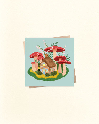Front of square greeting card featuring a whimsical illustration of a cottage surrounded by mushrooms, paired with a brown kraft envelope. 10cm x 10cm card by The Bubbly Type – Australian made and blank inside. Perfect for fairytale lovers, nature fans, or a magical little note.