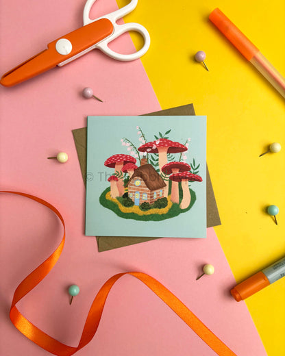 A square greeting card featuring a whimsical mushroom house illustration, styled in a flat-lay with an orange ribbon, scissors, pins, and markers on a split pink and yellow background.
