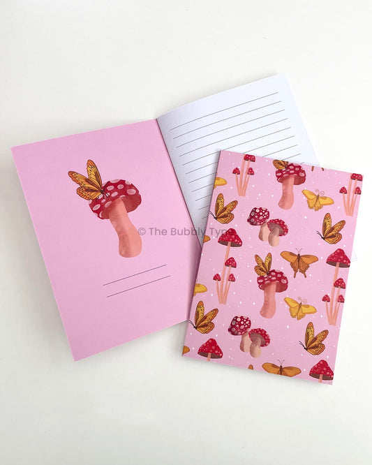 A5 notebook titled Butterflies and Mushrooms featuring a whimsical pattern design on the cover with butter yellow butterflies in various flight poses scattered among red-capped mushrooms of different shapes and sizes. The notebook is displayed open to show the inner front cover with an illustration of a red-capped mushroom topped by a yellow butterfly, alongside the first white lined page.