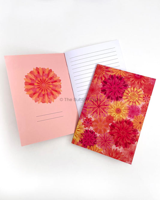 A5 notebook titled Floral Burst featuring a vibrant watercolour-style floral cover design with overlapping flowers in warm colours including deep pinks, reds, yellows, and oranges. The notebook is displayed with an open copy showing the inner front cover, which includes a pink and orange flower illustration, and the first white lined page.