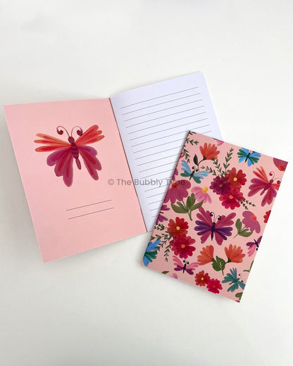 A5 notebook titled Butterfly Garden with a vibrant watercolor-style cover design featuring flowers, foliage, and butterflies in bright colours like blue, red, pink, orange, and green. The notebook is placed on an open copy of itself, showing the inner front cover with a pink butterfly illustration and the first lined page.