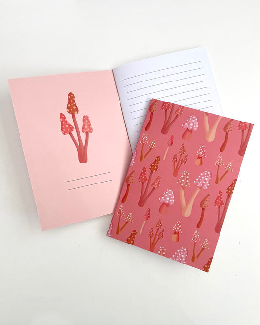 A5 notebook titled Boho Mushrooms with a watercolor-style cover featuring neutral-toned mushrooms and foliage. The notebook is placed on an open copy of itself, showing the inner front cover with a mushroom illustration and the first lined page.