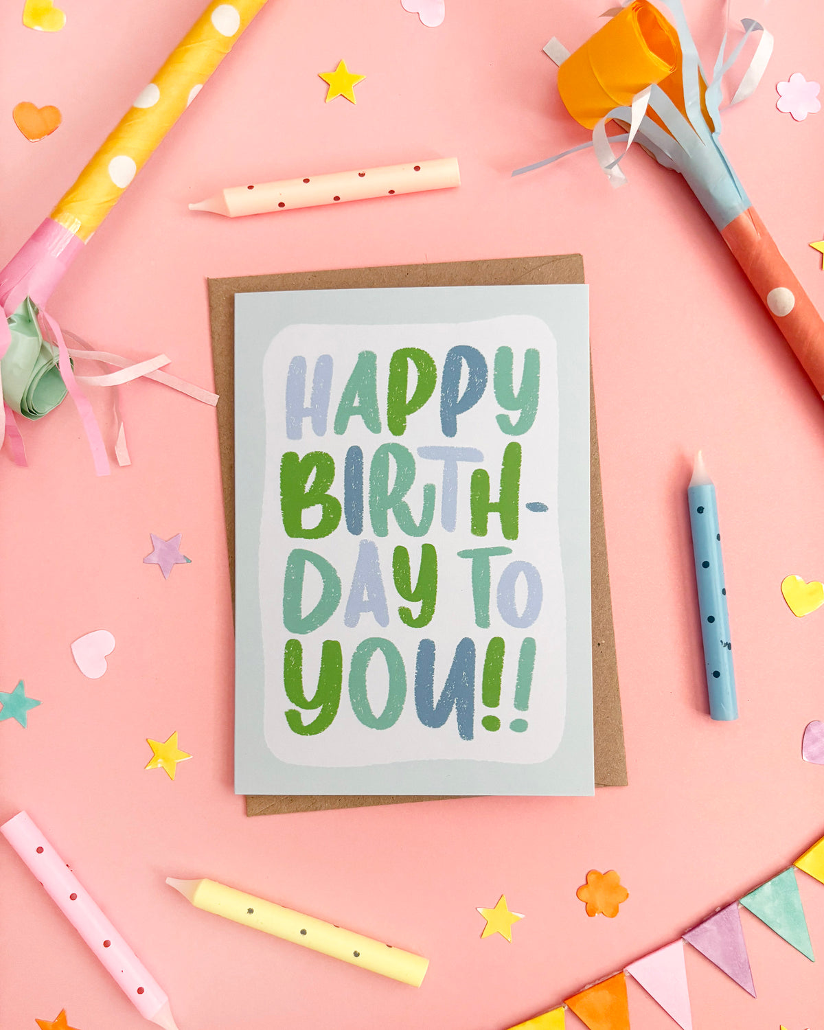 A birthday card with the words 'HAPPY BIRTHDAY TO YOU!!' in playful green and blue lettering, placed on a pink background with colourful party props including candles, streamers, confetti, and bunting.