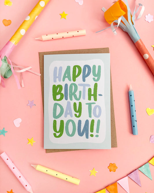 A birthday card with the words 'HAPPY BIRTHDAY TO YOU!!' in playful green and blue lettering, placed on a pink background with colourful party props including candles, streamers, confetti, and bunting.