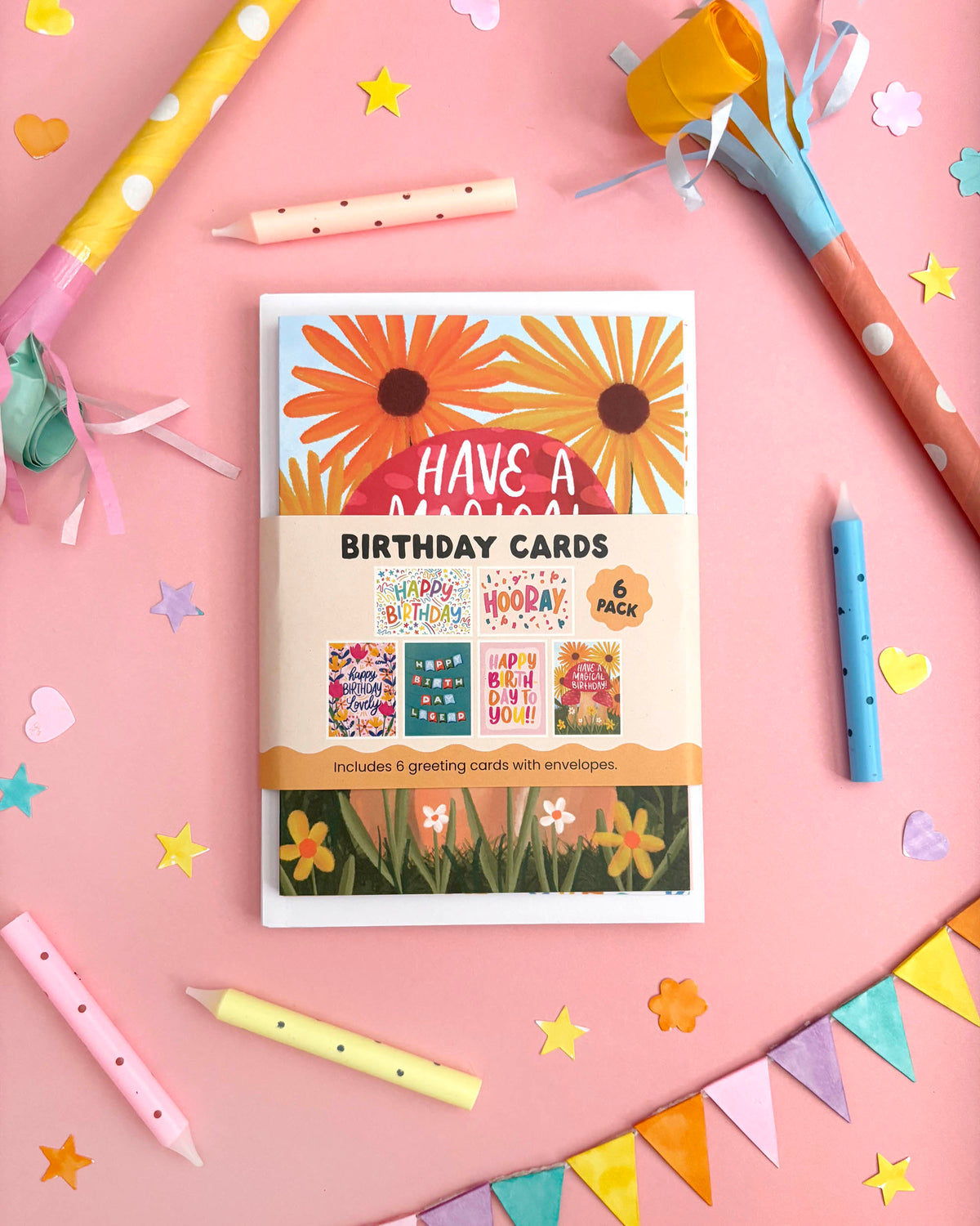 A boxed set of birthday cards displayed on a pink background with party-themed props including colourful candles, streamers, star confetti, and bunting. The front of the box shows large orange flowers and the text 'HAVE A MAGICAL BIRTHDAY CARDS' with smaller sample card designs visible inside.
