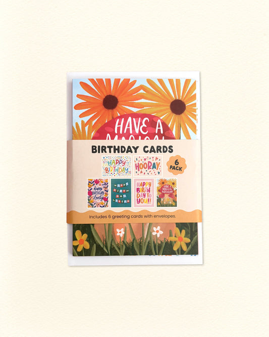 A 6-pack of 'Have a Magical Birthday Cards' in floral envelopes, featuring a white package with orange daisy illustrations and previews of colourful cards inside, set against a warm cream background.