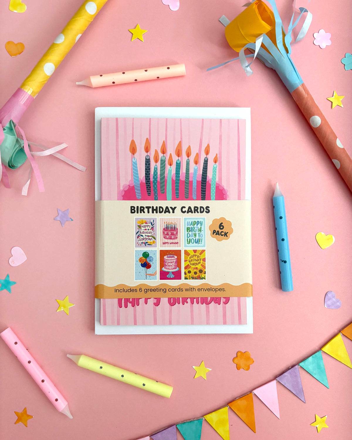 A boxed set of birthday cards arranged on a pink surface with party-themed props including candles, star confetti, paper party hats, and bunting. The front of the box shows the title 'BIRTHDAY CARDS' and a sample card design with lit candles and 'Happy Birthday!'.