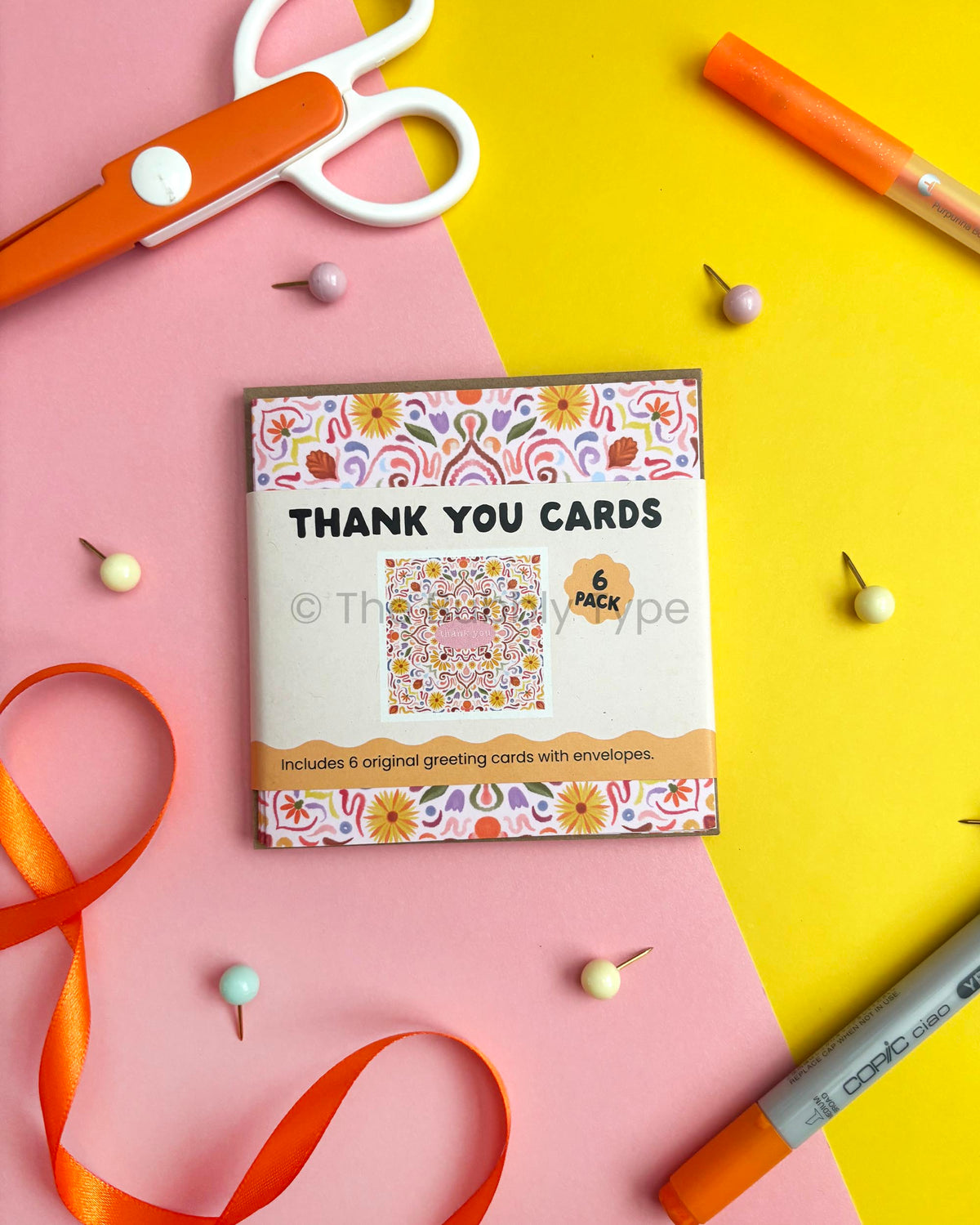 A 6-pack of thank you cards with colourful floral-patterned envelopes, displayed on a split pink and yellow background with craft props like scissors, pins, ribbon, and markers.