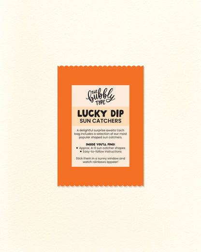 Photo of bright orange scalloped-edge lucky dip bag with a “Sun Catcher Lucky Dip” label listing what's included. Surprise pack of rainbow-making window decals from The Bubbly Type – a joyful gift idea for indecisive shoppers and lovers of light-filled decor.
