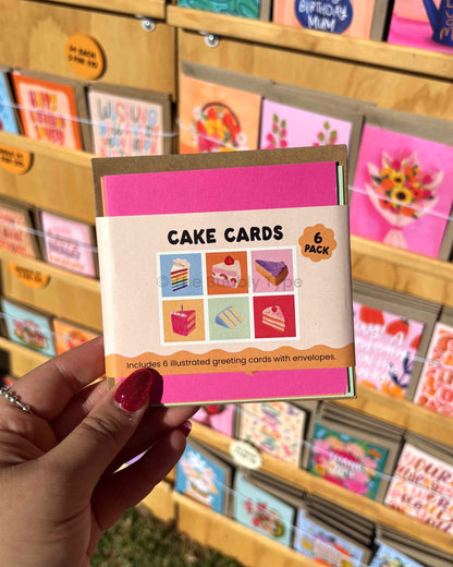 Hand holding a pack of cake-themed greeting cards with a store display in the background.