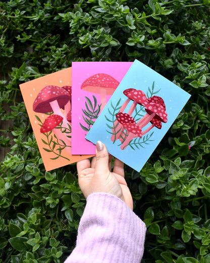 Three colourful greeting cards with mushroom designs held by a hand against a green leafy background.