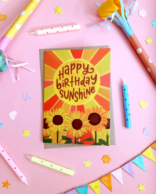 A birthday card with a sunburst design and three sunflowers, reading 'Happy Birthday Sunshine,' styled on a pink background with party candles, streamers, and star confetti.