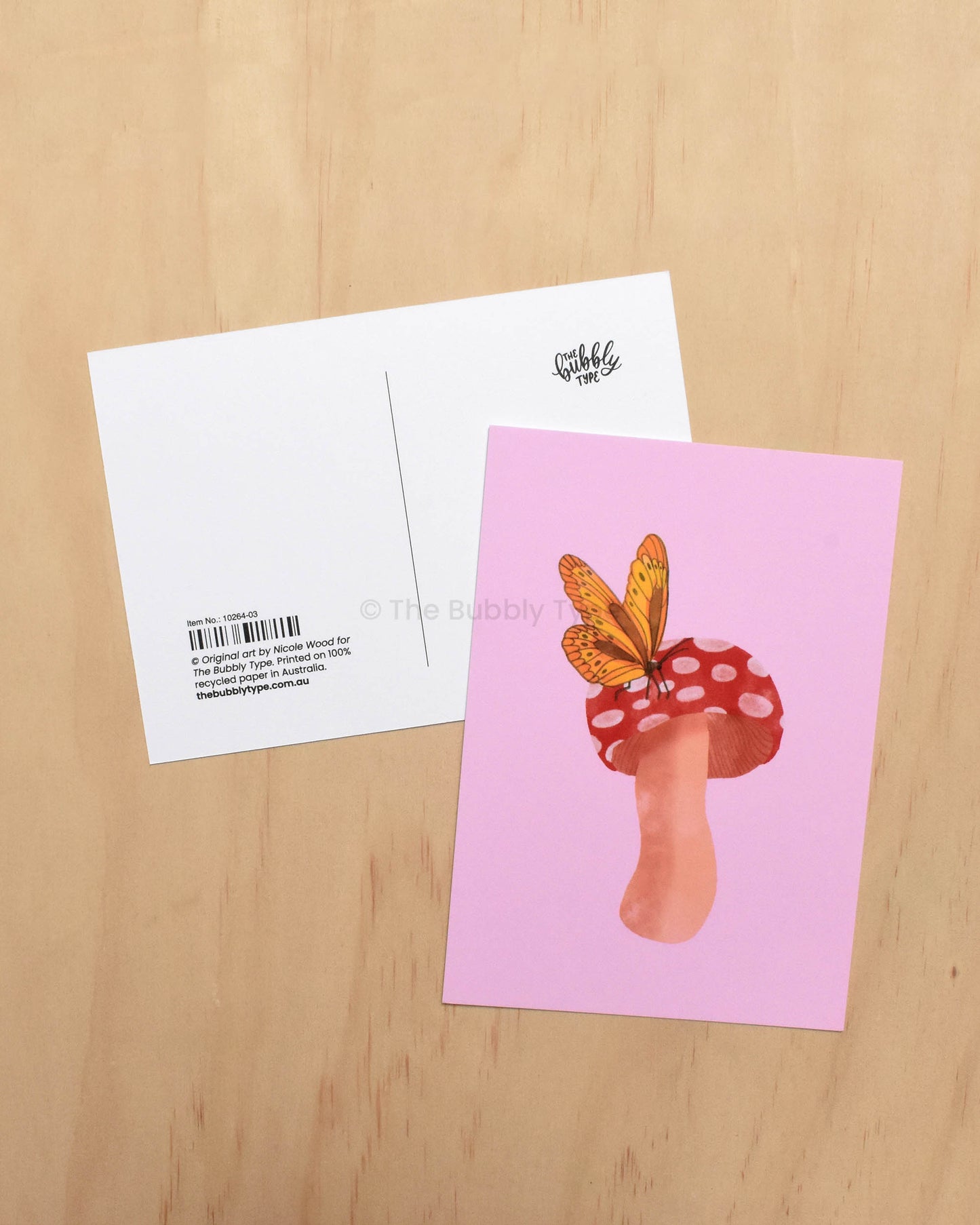 Postcard with a mushroom and butterfly design on a pink background, placed on a wooden surface.