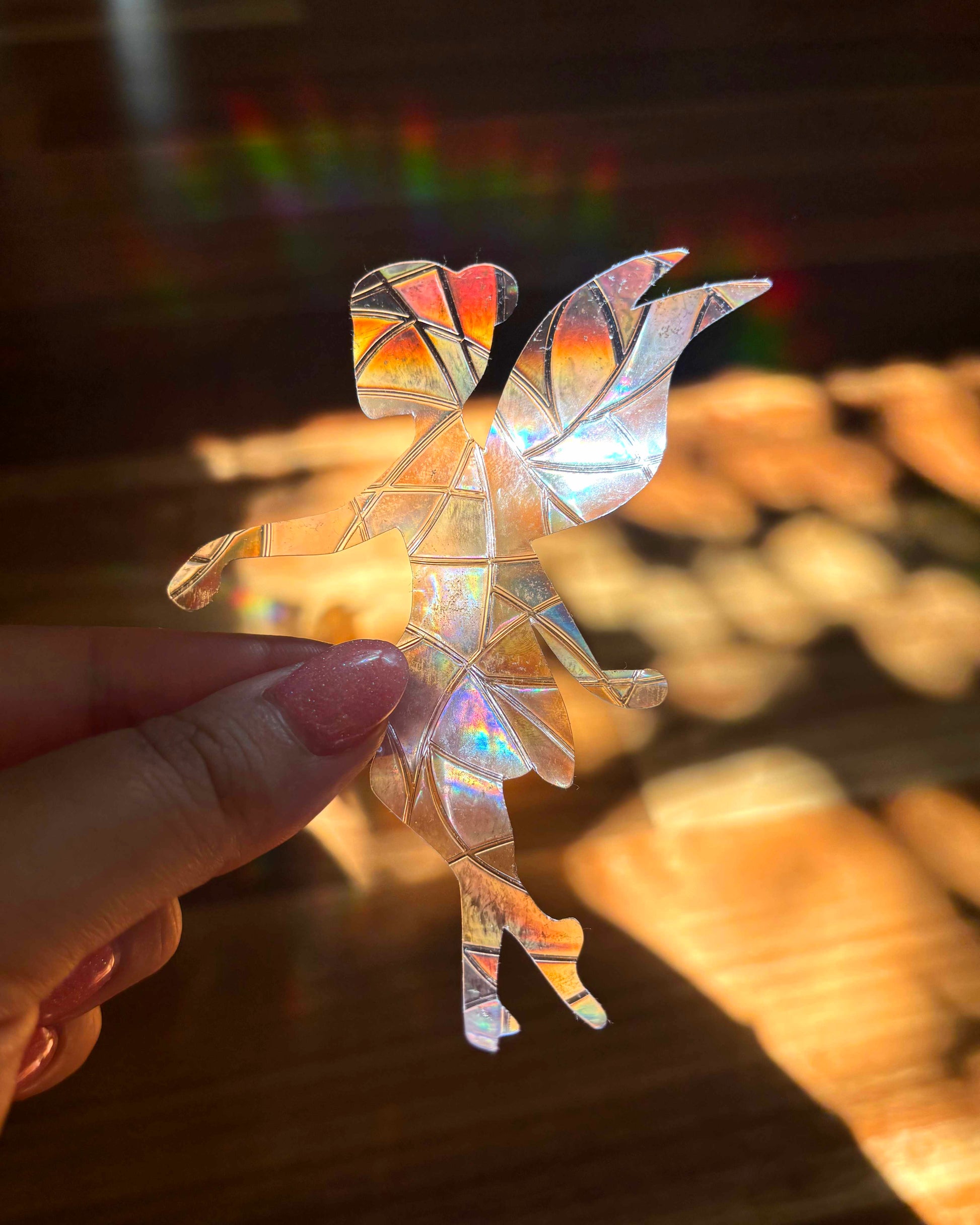 A hand holding an iridescent fairy-shaped sun catcher sticker with a background of rainbow lights.