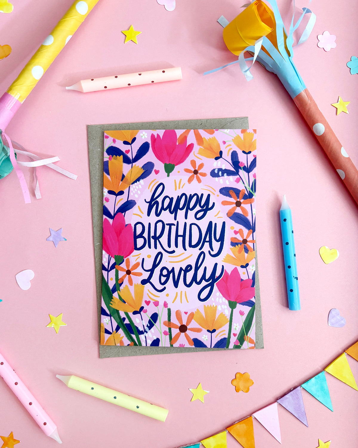 A floral birthday card with the words 'happy BIRTHDAY Lovely' in navy script, surrounded by colourful party props on a pink background.