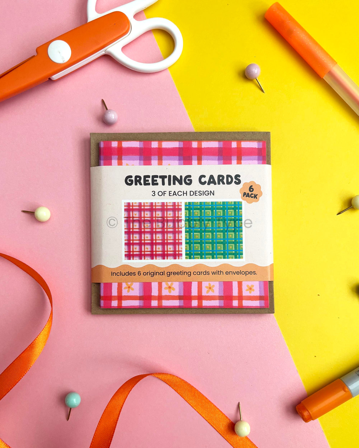 A flat-lay of a 6-pack of greeting cards on a split pink and yellow background with craft props like orange scissors, pins, and markers. The card packaging shows two plaid designs: pink and purple, plus green and blue.
