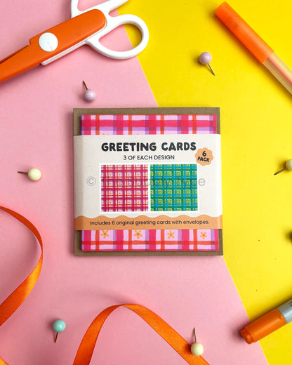 A flat-lay of a 6-pack of greeting cards on a split pink and yellow background with craft props like orange scissors, pins, and markers. The card packaging shows two plaid designs: pink and purple, plus green and blue.