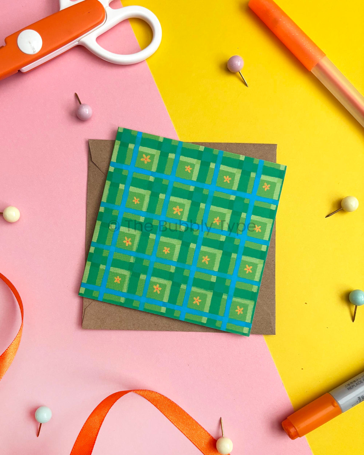 A square green patterned card or envelope with a blue grid and small yellow star motifs, placed on top of a brown kraft envelope. The item is styled in a flat lay on pink and yellow backgrounds with scissors, pins, ribbon, and markers around it.
