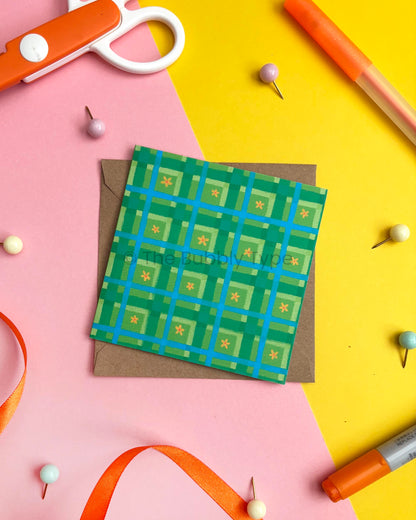 A square green patterned card or envelope with a blue grid and small yellow star motifs, placed on top of a brown kraft envelope. The item is styled in a flat lay on pink and yellow backgrounds with scissors, pins, ribbon, and markers around it.
