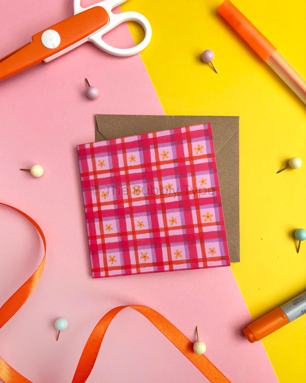 A square card with a pink, red, and purple plaid pattern decorated with small yellow flowers, placed on top of a brown envelope. The card is styled in a flat-lay arrangement with orange scissors, an orange marker, pins, and a ribbon on a split pink and yellow background.