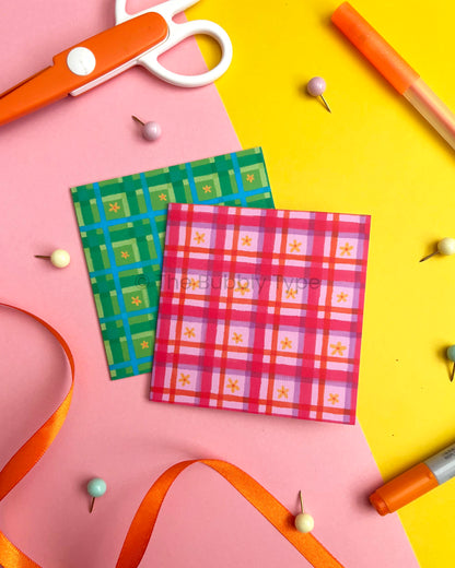 Two small square greeting cards with hand drawn gingham plaid patterns, one green and blue and one pink and purple, arranged on a pink and yellow background with craft props like scissors, pins, and ribbons.