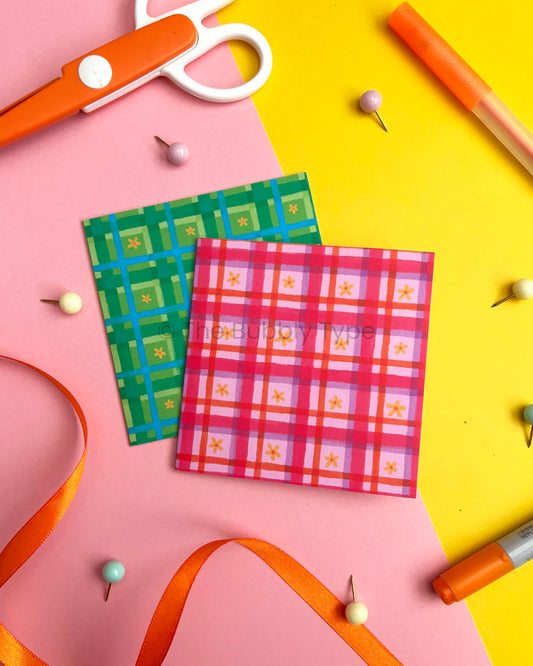 Two small square greeting cards with hand drawn gingham plaid patterns, one green and blue and one pink and purple, arranged on a pink and yellow background with craft props like scissors, pins, and ribbons.