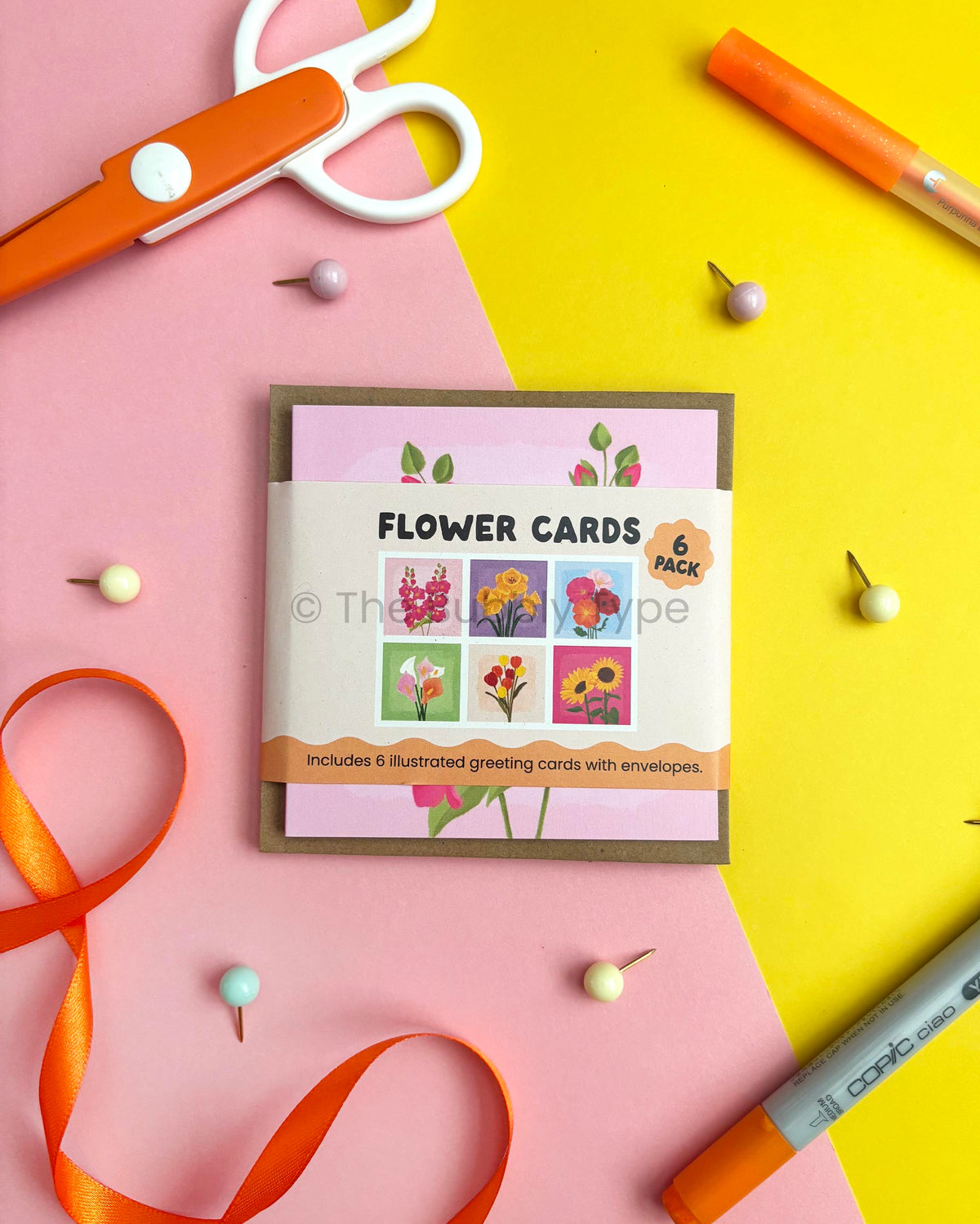 A 6-pack of illustrated flower greeting cards in a square envelope, styled in a flat-lay with orange scissors, a marker, a ribbon, and pins on a pink and yellow background.