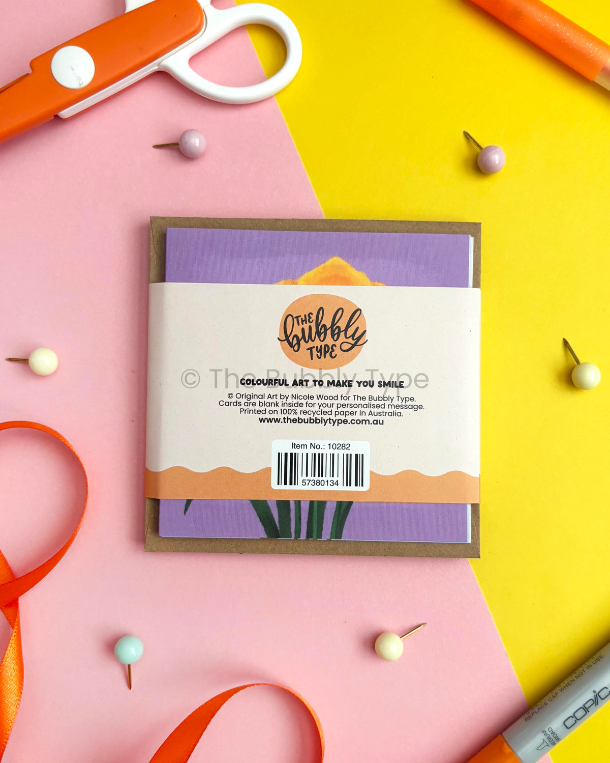 A square craft card or art print package from The Bubbly Type, featuring a purple and lavender design with a kraft paper border, displayed on a split pink and yellow background with craft props like scissors, beads, and ribbon.