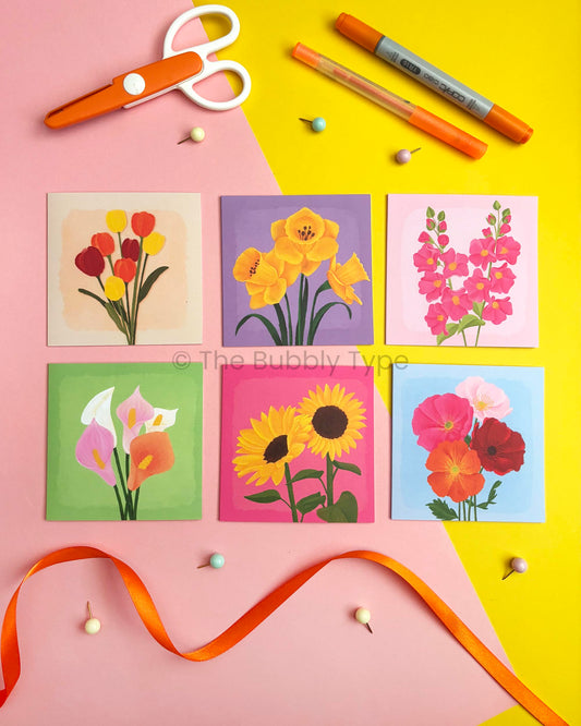 Six small square floral greeting cards arranged in a grid on a pink and yellow background, with orange scissors, markers, pins, and a ribbon around them. The cards show different flower illustrations: tulips, daffodils, hollyhocks, calla lilies, sunflowers, and poppies.
