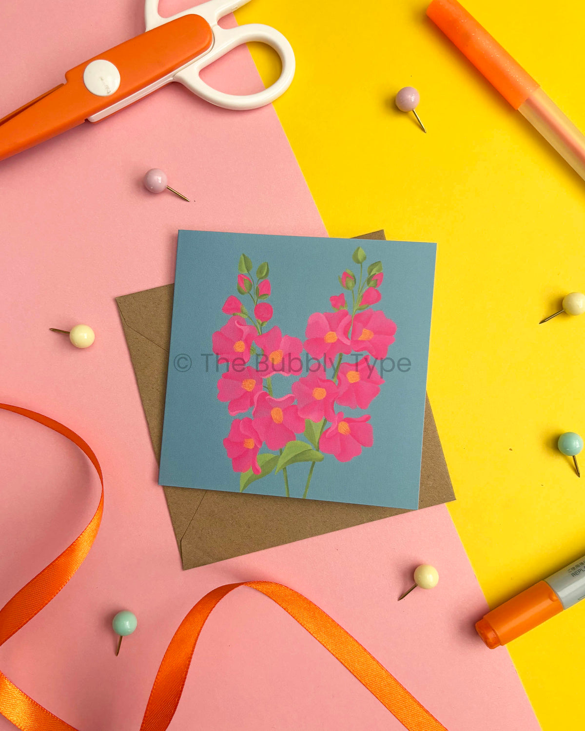 A square art print featuring a blue card with a pink flower illustration, styled in a flat-lay with a brown envelope, orange scissors, pins, ribbon, and markers on pink and yellow paper.