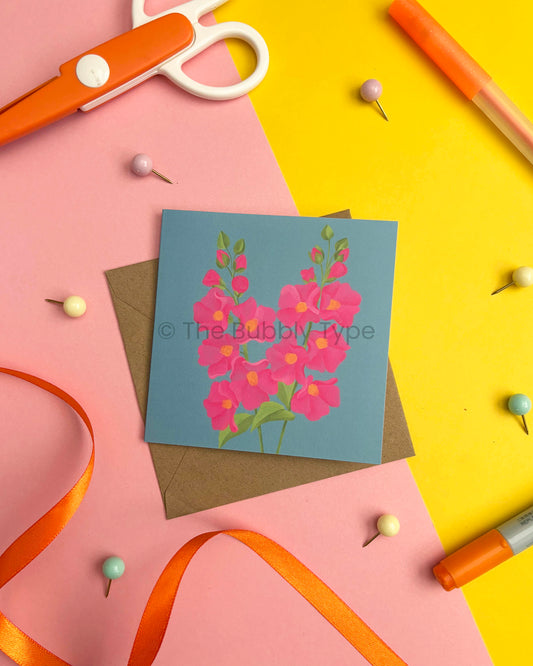 A square art print featuring a blue card with a pink flower illustration, styled in a flat-lay with a brown envelope, orange scissors, pins, ribbon, and markers on pink and yellow paper.