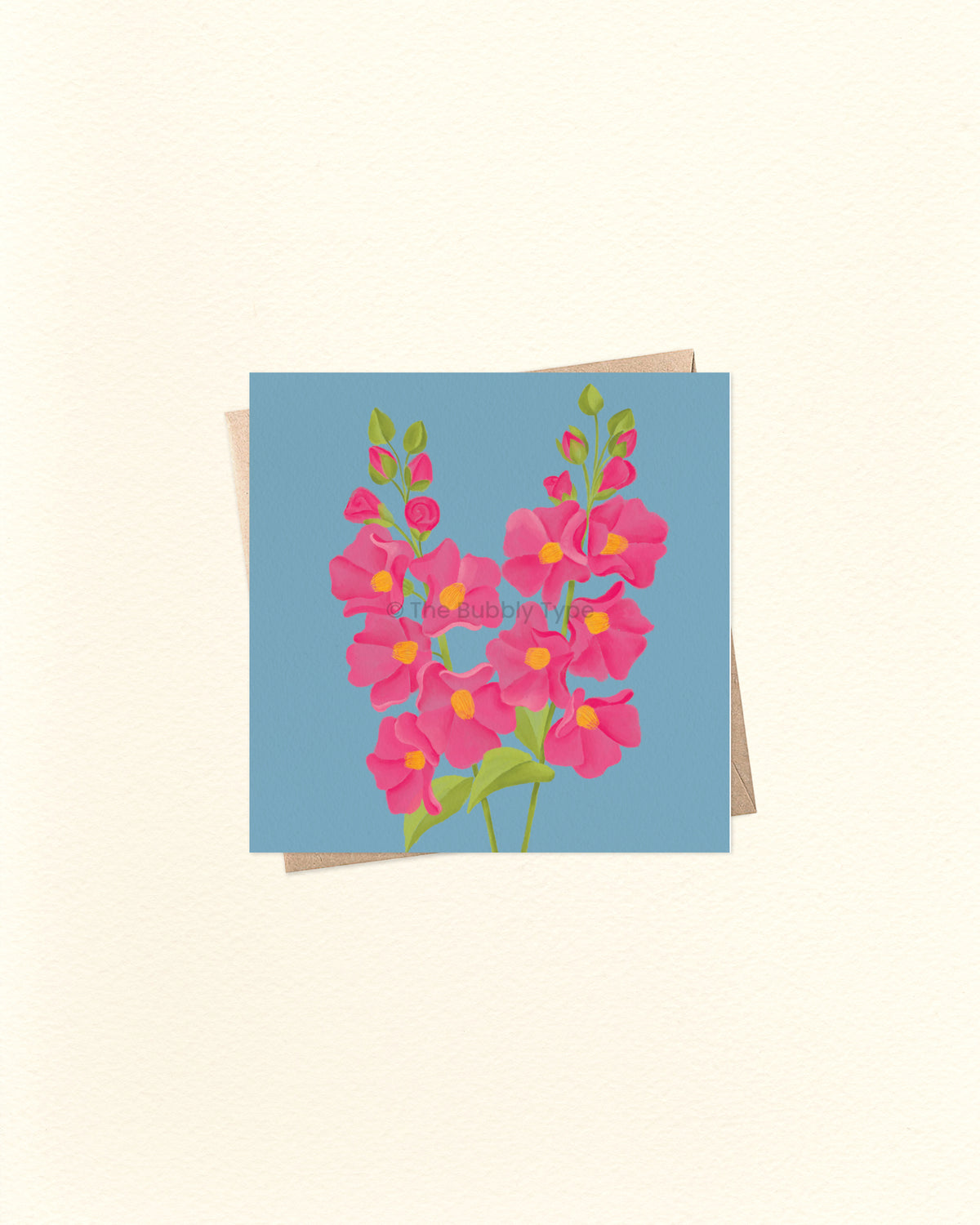 A square greeting card featuring a stylised floral bouquet in pink with yellow centres and green stems on a light blue background, shown with a kraft envelope behind it on a warm off-white surface.