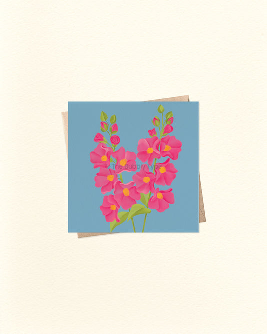 A square greeting card featuring a stylised floral bouquet in pink with yellow centres and green stems on a light blue background, shown with a kraft envelope behind it on a warm off-white surface.