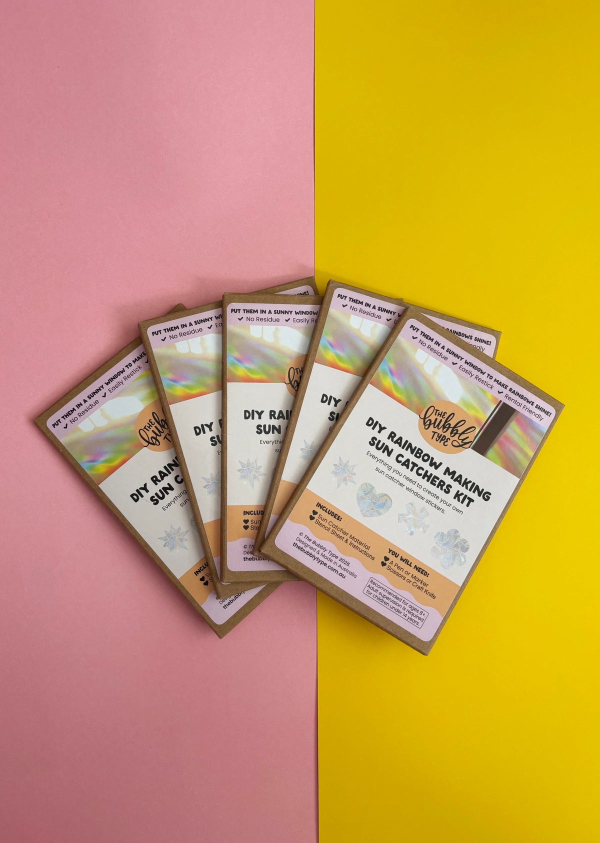 Five DIY Rainbow Making Sun Catchers Kits fanned out on a split pink and yellow background. 