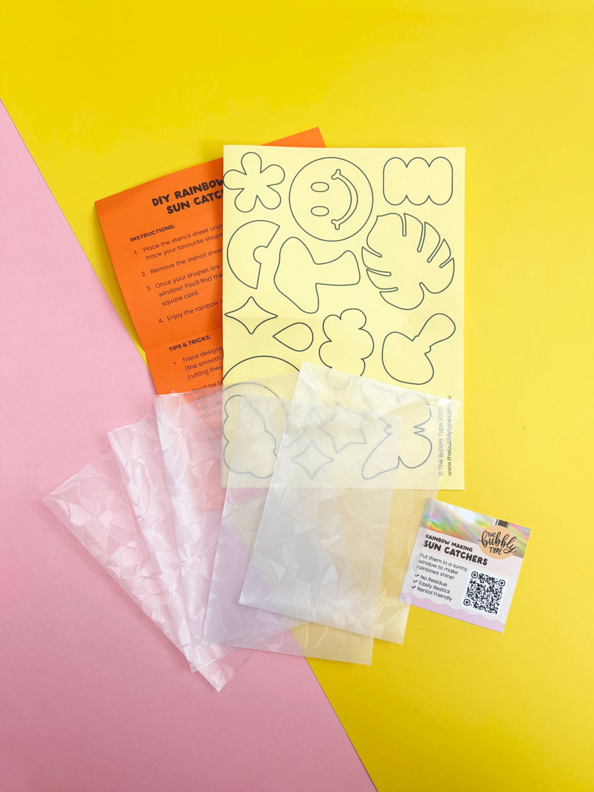 DIY rainbow sun catcher kit contents laid out on a diagonally split pink and yellow background, including clear sheets of sun catcher material, a yellow stencil sheet with outline templates, an orange DIY instruction card, and a small branded card with installation instructions.
