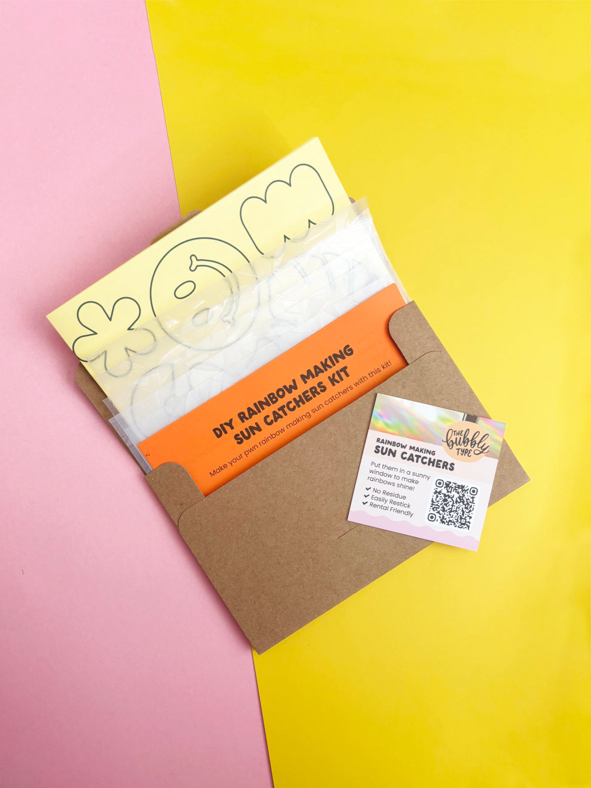 A DIY rainbow making sun catcher kit by The Bubbly Type, shown in a brown kraft envelope with printed templates and an orange instruction sheet, set against a split pink and yellow background.